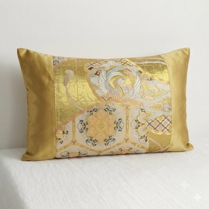 Puede incluir: Un cojín rectangular con un panel central de tela tejida en oro, crema y azul claro con un patrón de fénix y flores, flanqueado por paneles laterales de color oro mostaza sólido en los bordes izquierdo y derecho del cojín.