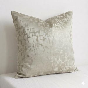 Può includere: Un cuscino decorativo quadrato in una palette di colori neutri. Il cuscino presenta un motivo strutturato e astratto nelle tonalità del beige e del grigio. Il cuscino è appoggiato su una superficie bianca, suggerendo un tema di arredamento per la casa.