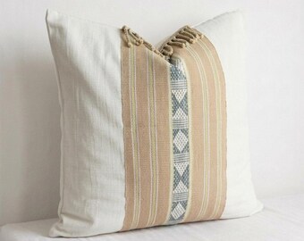 May include: A square throw pillow featuring a cream-colored fabric base with a central vertical panel. This panel displays tan, white, and light blue stripes alongside a geometric diamond pattern. The top edge includes decorative fringe.