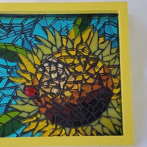 May include: A framed mosaic artwork depicting a sunflower with a ladybug on one of the petals. The sunflower is made of small pieces of glass in shades of yellow, brown, and green. The background is a blue and green mosaic.