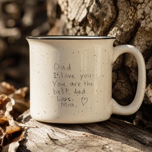 May include: A cream-colored ceramic mug with a black speckled pattern and a black rim. The mug features handwritten black text that reads: Dad I love you. You are the best dad Love, Mia, followed by a small hand-drawn heart symbol.