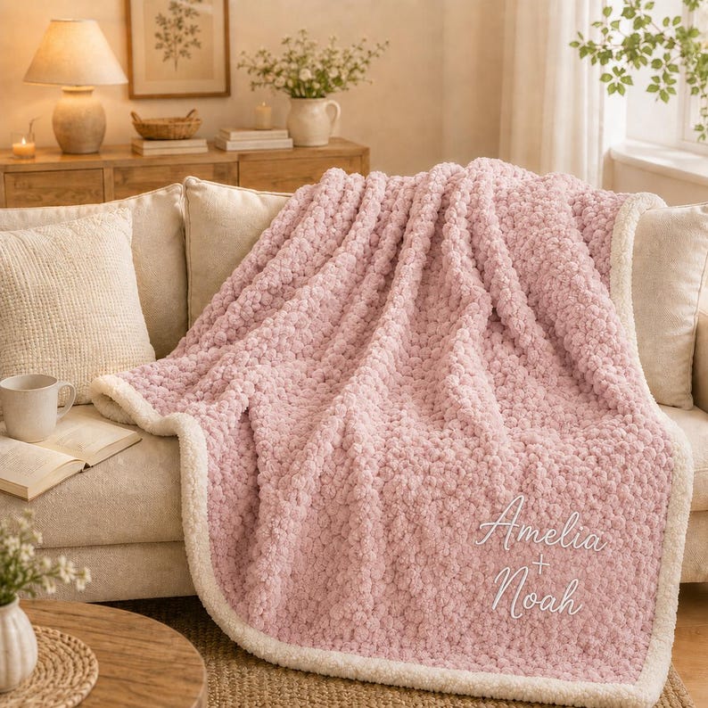 May include: A soft, textured light pink throw blanket with a white border. The blanket features the embroidered white text "Amelia + Noah" in a cursive script. The fabric has a plush, pom-pom-like appearance throughout the entire surface.