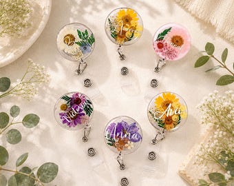 May include: A collection of clear, round badge reels with dried flowers and names. The flowers are yellow, pink, purple, and white. Each reel has a silver clip and a retractable cord. Names include Victoria, Ava, and Anna.