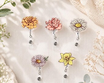 May include: Five floral-themed retractable badge holders. The designs include a marigold, a rose, a daisy, a pink daisy, and a daffodil. Each badge holder has a clear plastic base and a metal clip.