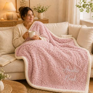 May include: A soft, textured, dusty pink throw blanket with a cream-coloured border. The blanket features the embroidered white text "Amelia + Noah" in a cursive script. The fabric has a plush, popcorn-like knit surface throughout the main body.