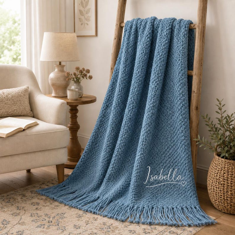 May include: A chunky knit throw blanket in a dusty blue color, featuring a textured pattern and long fringe along the bottom edge. The name Isabella is embroidered in white cursive script with a small heart icon on the lower right corner.
