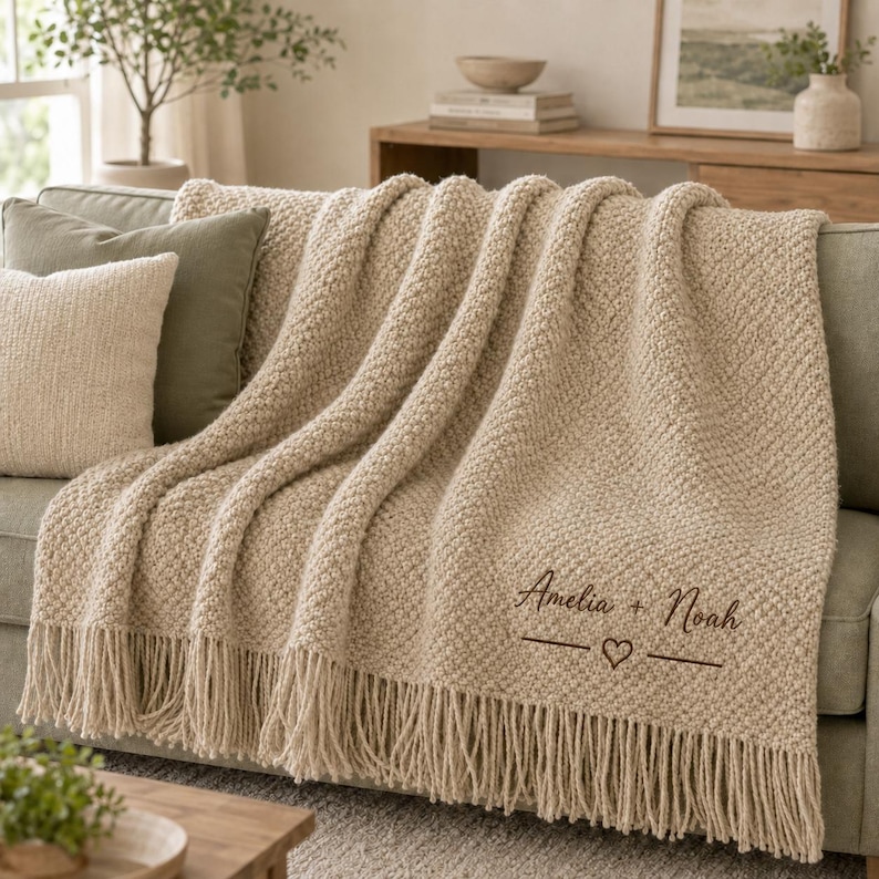 May include: A beige textured knit throw blanket with fringe trim, featuring the embroidered dark brown text "Amelia + Noah" above a horizontal line with a small heart symbol in the center, draped over a sofa with a matching beige pillow.