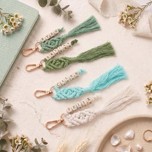 May include: Macrame keychains in various colors, including green, blue, and white, with gold-colored clasps. Each keychain features a tassel and wooden beads spelling out names like "Ryan," "Skylar," "Caleb," and "Felix." The keychains are arranged on a light-colored surface.