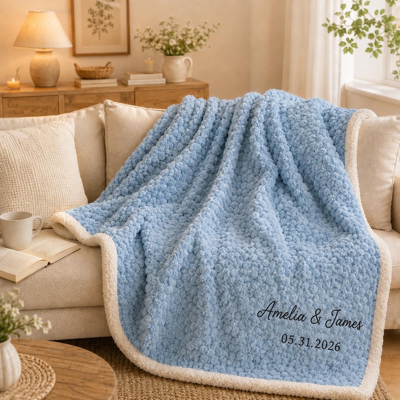 May include: A light blue textured throw blanket with a white sherpa lining. The blanket features embroidered black text that reads Amelia & James and the date 05.31.2026 in the bottom right corner. The fabric has a soft, raised, pom-pom-like texture.