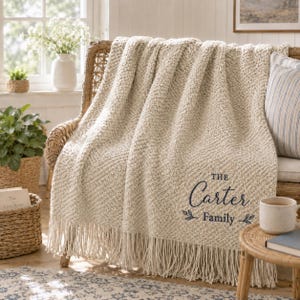 May include: A cream-colored knit throw blanket with a textured pattern and long fringe along the bottom edge. The blanket features dark blue embroidered text that reads "THE Carter Family" with small leaf accents on either side of the word Family.