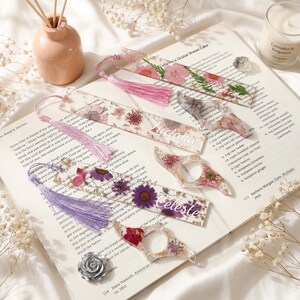 Könnte beinhalten: Transparente Lesezeichen und Buchseitenhalter aus Harz mit getrockneten Blumen und Goldflocken. Die Lesezeichen haben rosa, lila und weiße Quasten. Ein Lesezeichen hat den Namen "Luciana" und ein anderes den Namen "Celeste".