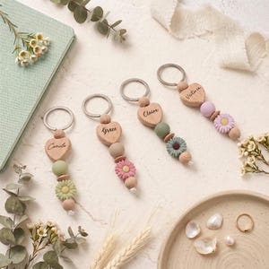May include: Four personalized keychains with wooden heart pendants engraved with names. Each keychain features a variety of beads in pastel colors, including pink, green, and lavender, and a flower charm. The keychains are arranged on a white surface.