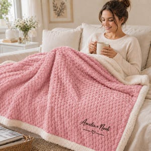 May include: A soft, textured pink blanket with a white sherpa-style lining. The blanket features the embroidered text "Amelia + Noah" above a small heart symbol and two horizontal lines, positioned in the lower right corner of the pink fabric.