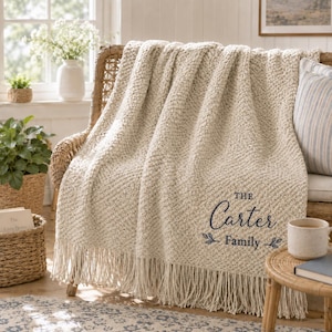 May include: A cream-colored knit throw blanket with a textured pattern and long fringe along the bottom edge. The blanket features dark blue embroidered text that reads "THE Carter Family" with small leaf accents on either side of the word Family.