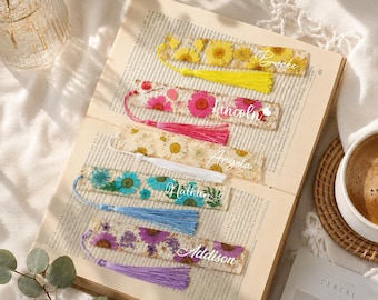 Puede incluir: Un conjunto de marcapáginas de resina hechos a mano, cada uno con flores secas y una borla. Los marcapáginas rectangulares y transparentes tienen nombres como "Brooke", "Lincoln", "Angela", "Mathias" y "Addison". Las borlas son amarillas, rosas, blancas, azules y moradas.