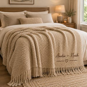 Peut inclure: Un plaid en tricot texturé beige doté d'une frange décorative sur le bord inférieur. Le plaid est personnalisé avec le texte brodé marron foncé Amelia + Noah, positionné au-dessus d'un petit symbole de cœur et de lignes horizontales.