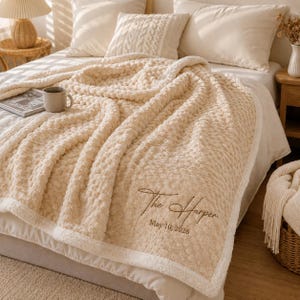 May include: A cream-coloured textured throw blanket with a soft, bumpy surface and a white border. The blanket features dark brown embroidered text that reads The Harper and the date May 10, 2026, positioned in the lower right corner of the fabric.