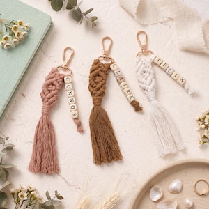 Pode incluir: Três chaveiros de macramé em tons de rosa, castanho e branco. Cada chaveiro apresenta um fecho dourado, contas de madeira com um nome e uma borla. Os chaveiros são exibidos em uma superfície clara com detalhes florais.