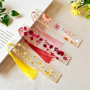 Peut inclure: Quatre marque-pages personnalisés avec des fleurs pressées et des accents de feuilles d'or. Les marque-pages sont transparents et ont un pompon attaché. Les noms sur les marque-pages sont Vivienne, Adelaide, Beckham et un quatrième nom qui n'est pas clair.