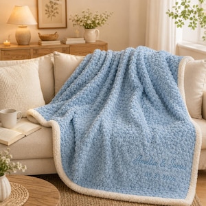 May include: A light blue textured throw blanket with a white sherpa lining. The blanket features embroidered text in a cursive font that reads Amelia & James followed by the date 05.31.2026 in a simple sans-serif font on the bottom right corner.