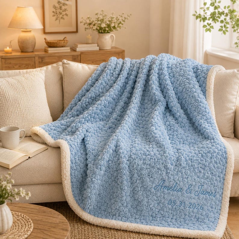 May include: A light blue textured throw blanket with a white sherpa lining. The blanket features embroidered text in a cursive font that reads Amelia & James followed by the date 05.31.2026 in a simple sans-serif font on the bottom right corner.