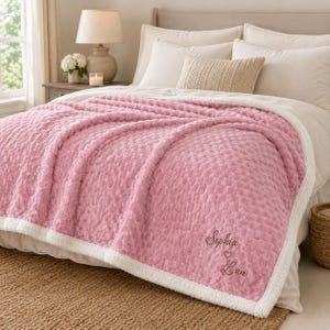 May include: A soft, textured pink blanket with a white sherpa lining. The blanket features embroidered text that reads Sophia, a small heart symbol, and Liam in a dark thread. The fabric has a raised, grid-like pattern across the surface.
