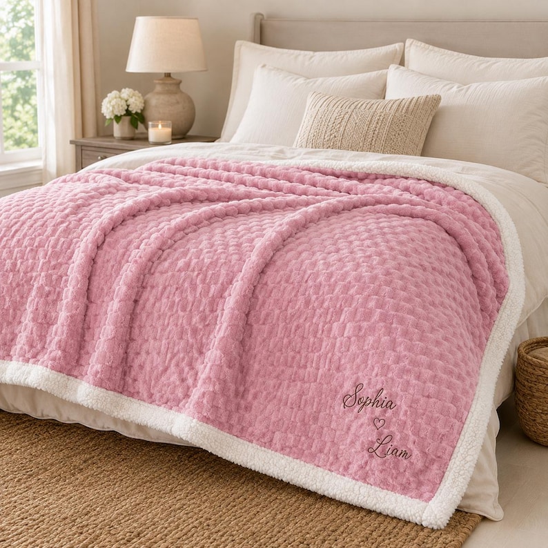 May include: A soft, textured pink blanket with a white sherpa lining. The blanket features embroidered text that reads Sophia, a small heart symbol, and Liam in a dark thread. The fabric has a raised, grid-like pattern across the surface.