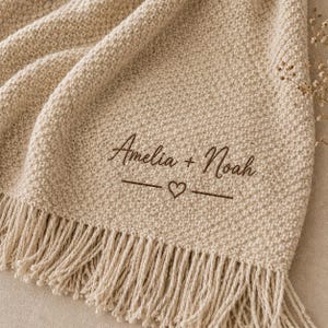May include: A beige knit throw blanket featuring a textured pattern and fringe trim along the bottom edge. The fabric is embroidered with the dark brown text Amelia + Noah, positioned above a horizontal line with a small heart in the center.