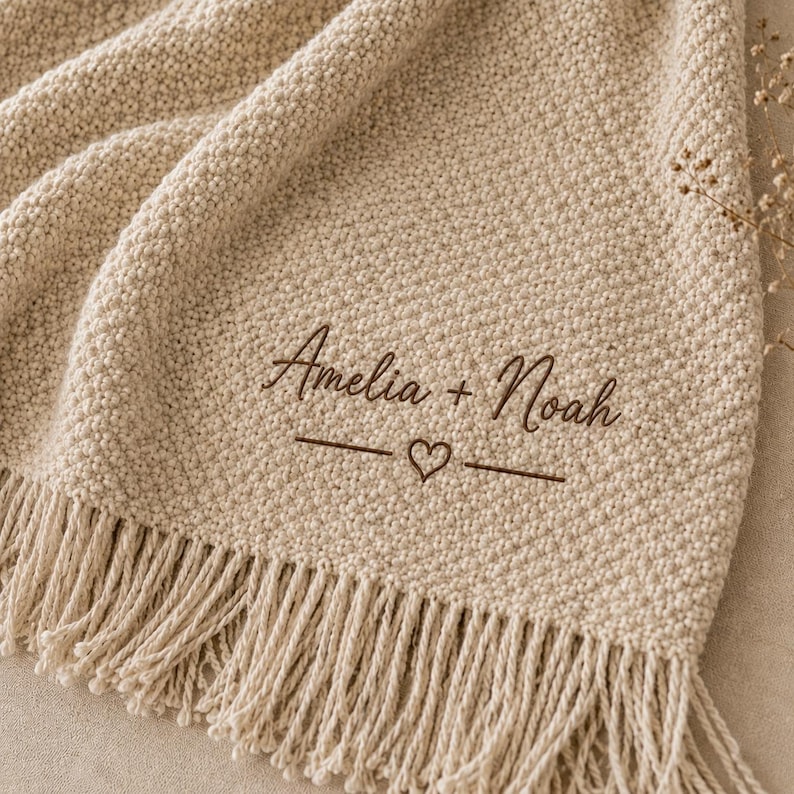May include: A beige knit throw blanket featuring a textured pattern and fringe trim along the bottom edge. The fabric is embroidered with the dark brown text Amelia + Noah, positioned above a horizontal line with a small heart in the center.