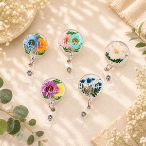 Può includere: Cinque porta badge rotondi e trasparenti con fiori pressati colorati e nomi in corsivo. I fiori includono fiori blu, arancioni, viola, gialli e bianchi. Ogni porta badge ha un cordino retrattile e una clip di plastica.