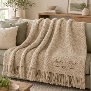 May include: A beige textured knit throw blanket with fringe trim, featuring the embroidered dark brown text "Amelia + Noah" above a horizontal line with a small heart symbol in the center, draped over a sofa with a matching beige pillow.