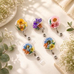 Può includere: Cinque porta badge rotondi e trasparenti con fiori secchi colorati e nomi in corsivo: Angela, Victoria, Hannah, Ava e Sophie. Ogni rotolo ha un cordino retrattile e una clip in metallo per attaccarlo ai vestiti.