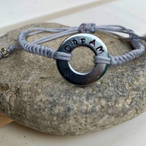 May include: A gray bracelet with a silver charm that says "DREAM" on it. The bracelet is made of a braided cord and has a gold bead on each end.