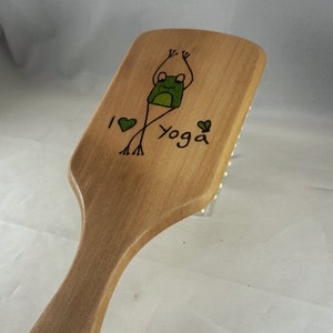 Può includere: Una spazzola per capelli in legno con un'illustrazione di rana verde e il testo "Amo lo yoga" sul retro.