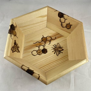 May include: A hexagonal wooden bowl with a natural wood finish. The bowl is decorated with a wood-burned design of honeycombs and bees.