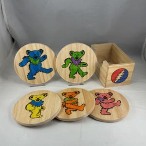 May include: A set of five circular wooden coasters featuring hand-drawn dancing bears in blue, green, yellow, orange, and pink, accompanied by a matching wooden storage box with a circular red, white, and blue lightning bolt emblem on the front.