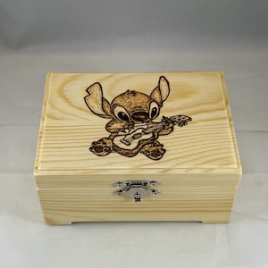 Puede incluir: Una caja de madera de color claro con tapa con bisagras y cierre plateado. La parte superior de la caja presenta una imagen detallada, grabada en madera, de un personaje de dibujos animados tocando la guitarra. La caja es rectangular y tiene un acabado natural simple.