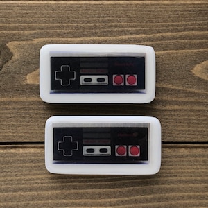 May include: Two white rectangular resin keychains with a black and white image of a classic video game controller. The controller has a red cross, two gray buttons, and two red buttons.