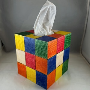 Puede incluir: Un dispensador de pañuelos de madera de colores con un diseño de cubo. La caja está hecha de cuadrados individuales en varios colores, incluyendo rojo, azul, verde, amarillo y naranja. Los pañuelos blancos son visibles en la parte superior.
