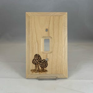May include: A light-colored wooden light switch cover plate featuring a rectangular center cutout and two screw holes. The bottom left corner displays a detailed wood-burned illustration of two morel mushrooms with textured caps and stems.