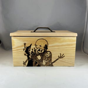 Può includere: Una scatola di legno con un coperchio incernierato e una maniglia in metallo. La scatola è decorata con un'immagine bruciata al legno di Jack Skellington e Sally di The Nightmare Before Christmas.