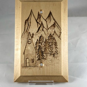 May include: A wooden light switch cover with a woodburned design of a cabin in the woods. The design features a small cabin with a window and a door, surrounded by trees and mountains.