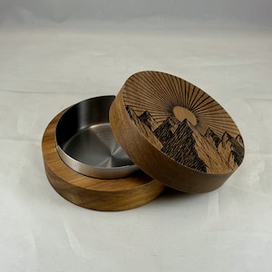 May include: A wooden box with a lid featuring a black and white mountain scene with a sun. The box has a stainless steel interior.