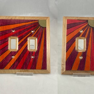 May include: A double light switch cover with a wood frame and a sunburst design in shades of red, orange, and purple.