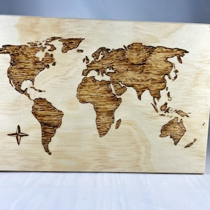 Puede incluir: Un mapa del mundo de madera, pirograbado sobre un panel de madera clara. Los continentes están representados en un diseño marrón quemado detallado. Una rosa de los vientos está en la esquina inferior izquierda. La veta de la madera es visible.