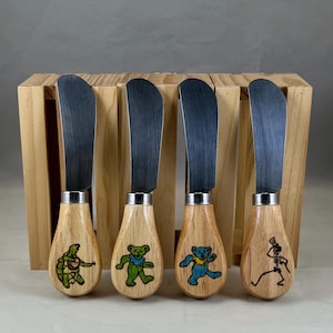 Puede incluir: Juego de cuatro cuchillos de mantequilla con mangos de madera y hojas de acero inoxidable. Cada mango presenta un gráfico colorido diferente: una tortuga tocando la guitarra, un oso verde, un oso azul y un esqueleto bailando. Los cuchillos se exhiben en una caja de madera.
