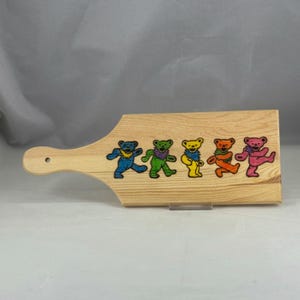 May include: A light-colored wooden cutting board with a handle. The board features five colorful dancing bear designs in blue, green, yellow, orange, and pink. The bears are in a row, adding a playful touch to the kitchen item.