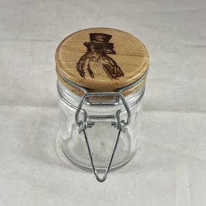 Puede incluir: Un tarro de vidrio transparente con una tapa de madera que presenta un diseño de pájaro con sombrero de copa y gafas quemado en madera. El tarro tiene un cierre de metal.