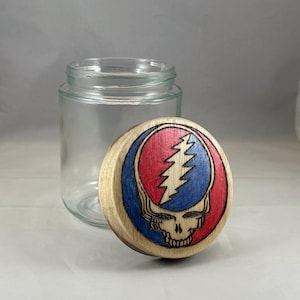 Puede incluir: Un tarro de vidrio transparente con una tapa de madera que presenta un diseño colorido. La tapa muestra una calavera con un rayo, sobre un fondo rojo y azul. El tarro está vacío y está junto a la tapa.