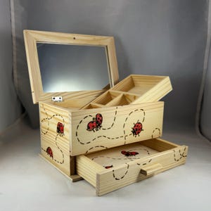 May include: Wooden jewelry box with a hinged mirror and two drawers. The box is decorated with red ladybug designs and dotted lines. The top section has compartments for organizing jewelry. The box is made of light-colored wood.